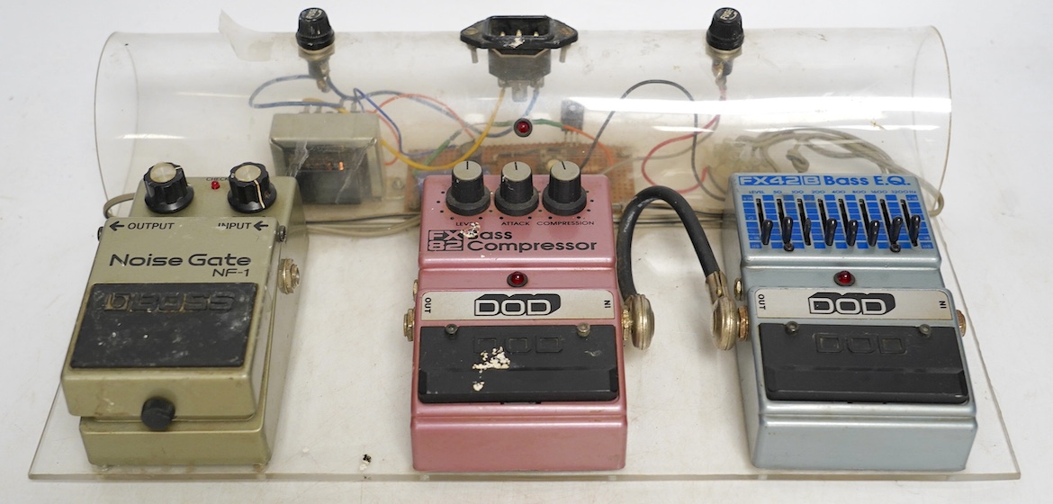 A bass guitar pedal board including; a DOD FX42 B bass EQ pedal, a DOD FX82 bass compressor and a Boss noise gate NF-1 pedal, mounted on a clear Perspex frame. Condition fair, some wear from use evident.